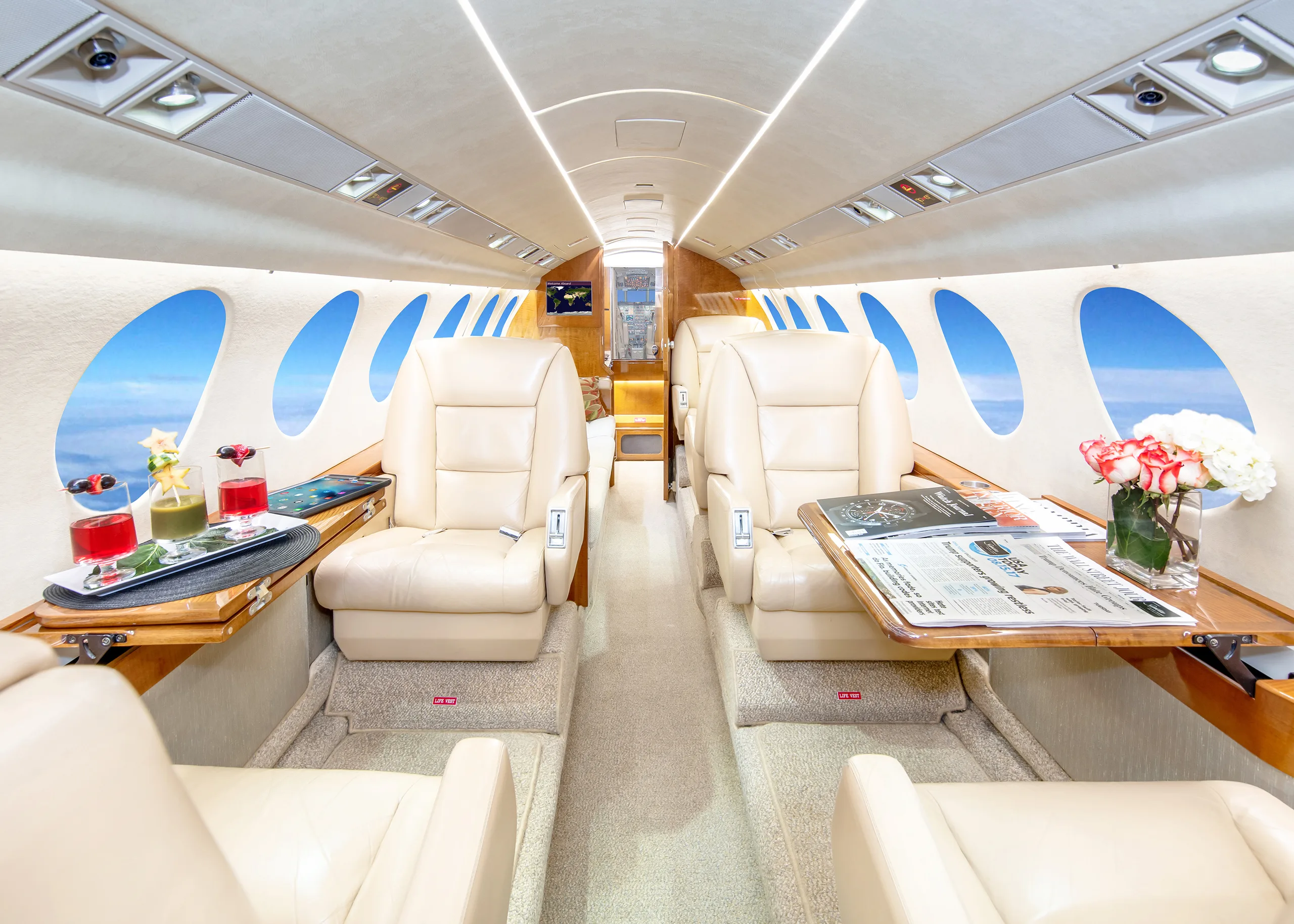 The elegant interior of a private jet cabin featuring beige leather seats, fold-out tables with drinks and a newspaper, and windows showing blue sky.