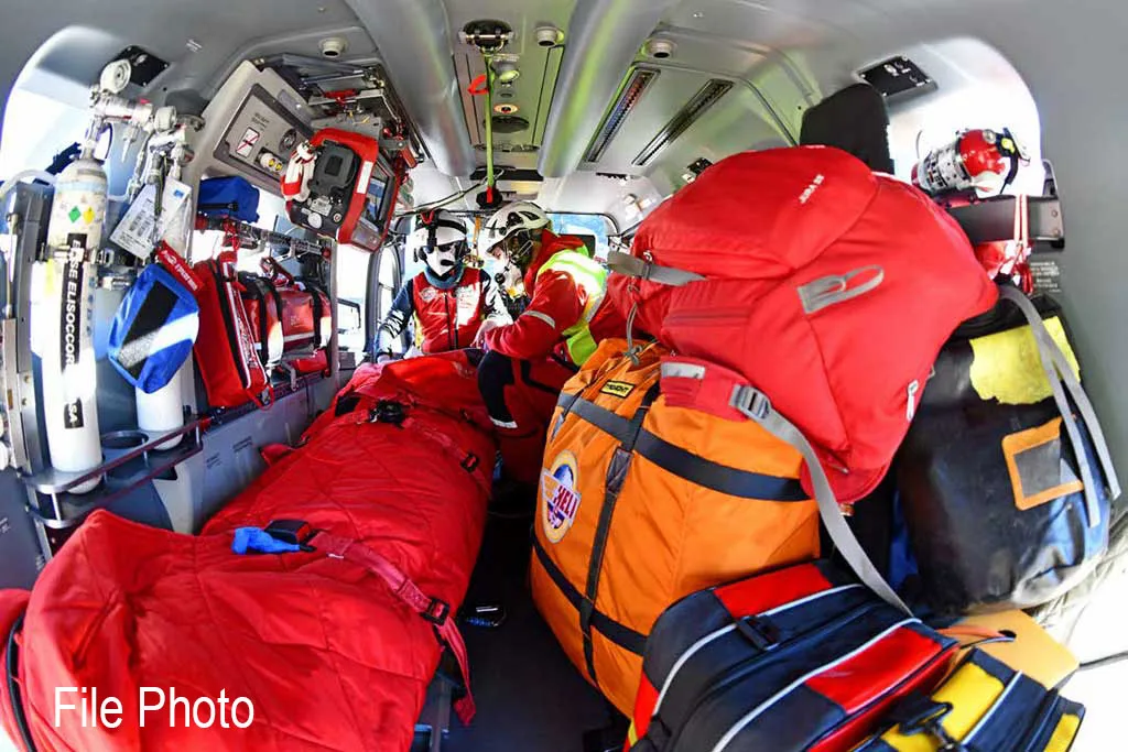 The interior of a helicopter is configured for emergency services, showing medical equipment, large bags, and two crew members in the cockpit area.