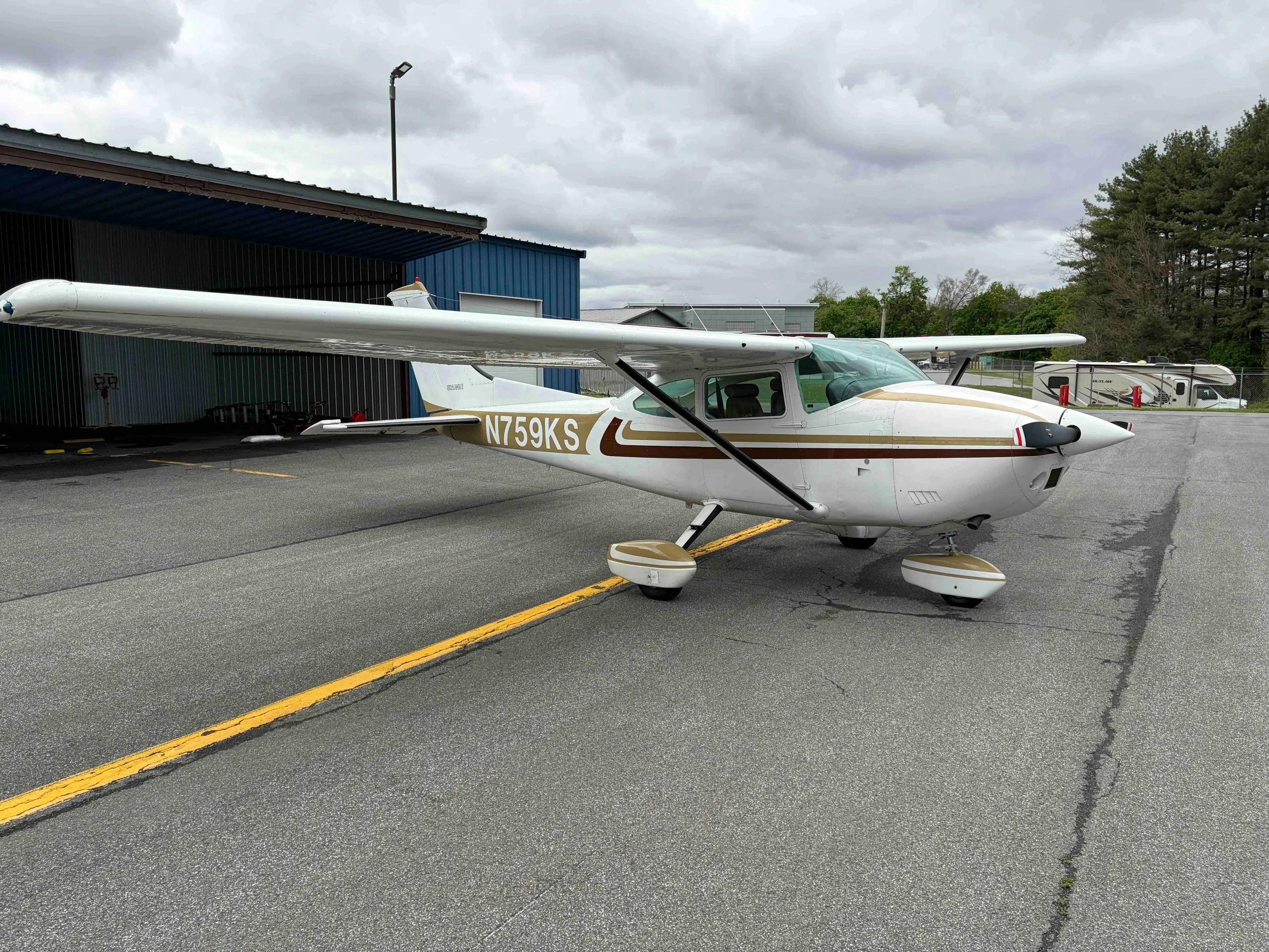 A front three-quarter view of the white Cessna 182 Skylane N759KS with gold and brown stripes, parked on an airport tarmac.