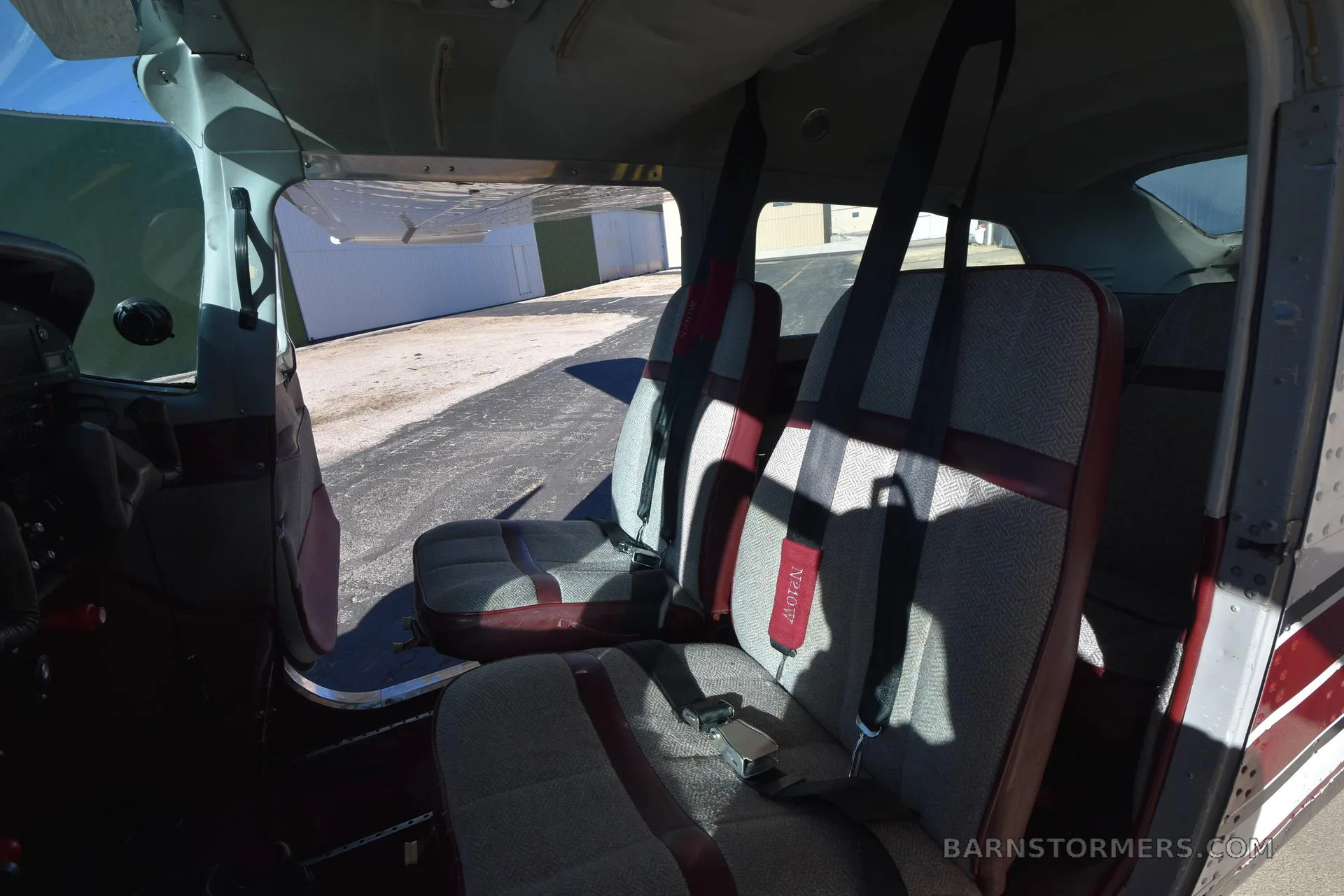 The interior cabin of a Cessna aircraft, showing two rows of grey and maroon fabric seats with seatbelts.