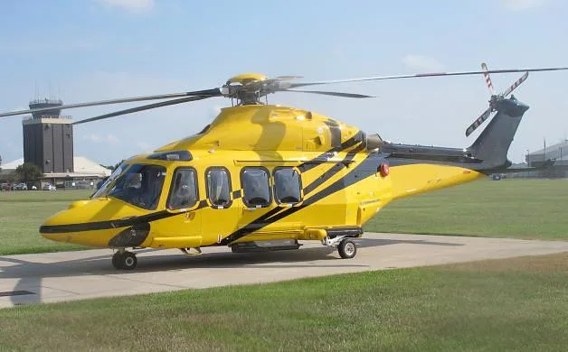 A yellow helicopter with black stripes parked on a tarmac with a control tower in the background.