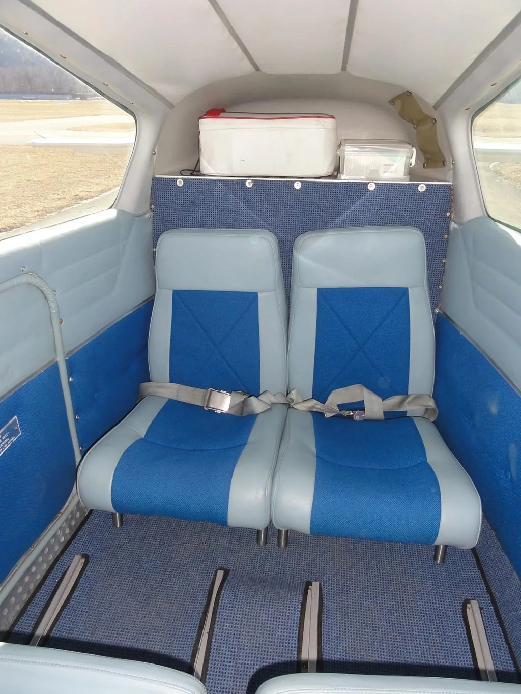 The aircraft's rear cabin with two blue and light grey seats, seatbelts, and a cooler on the shelf.