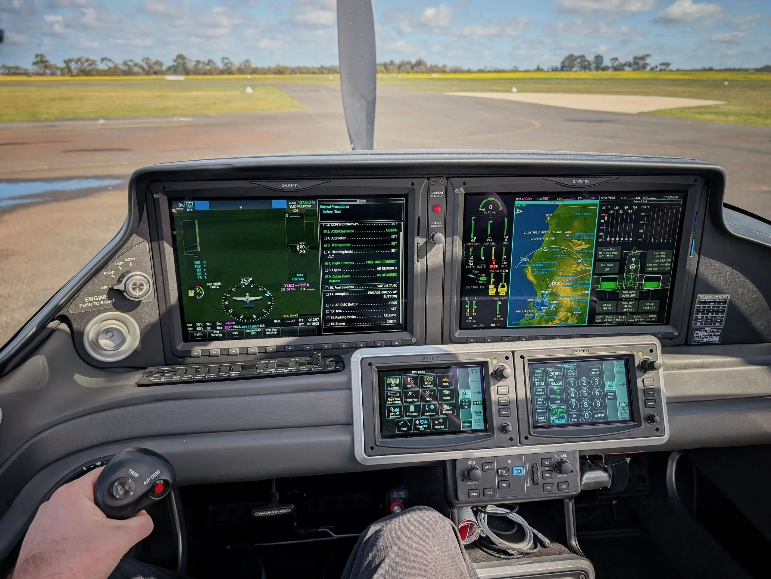 The cockpit of an aircraft with multiple Garmin glass displays and a control stick, looking out onto a runway.