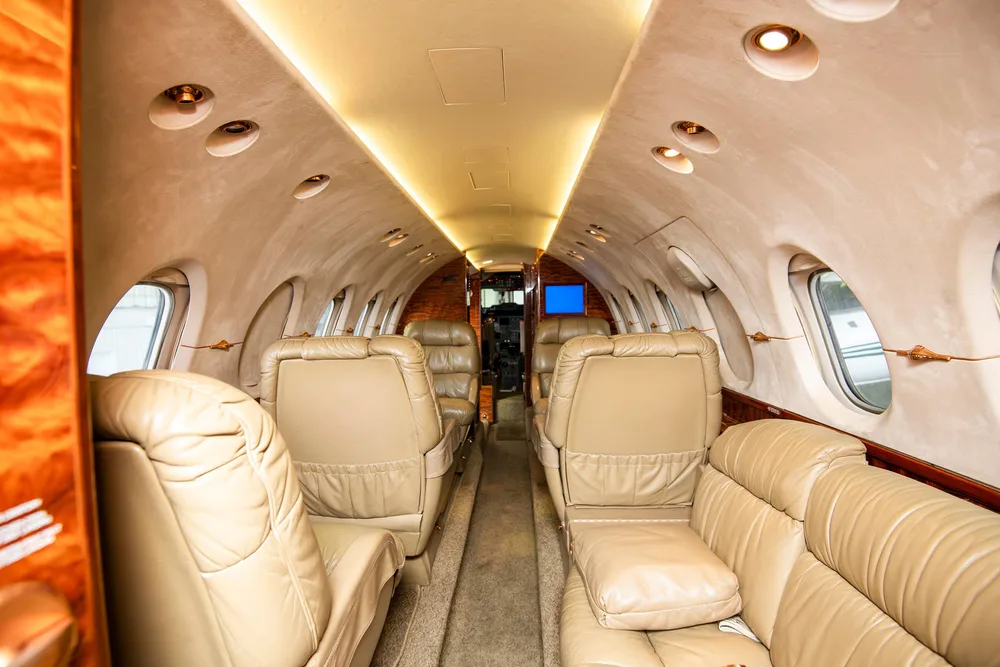 A view down the aisle of a private jet's cabin, showing multiple rows of tan leather seats and wood paneling.