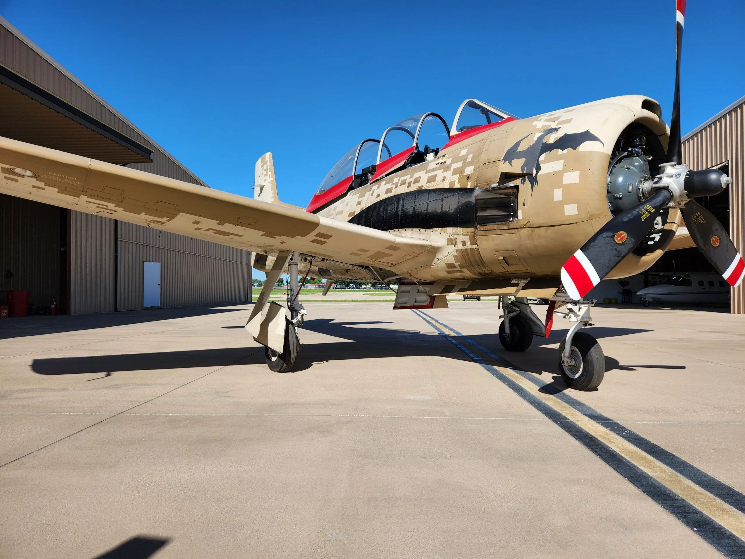 A T-28 Trojan aircraft with desert digital camouflage and red accents is parked on an airfield, showing its propeller and landing gear.