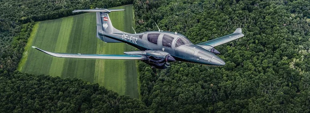 A gray twin-engine aircraft is in flight over a forest and field.