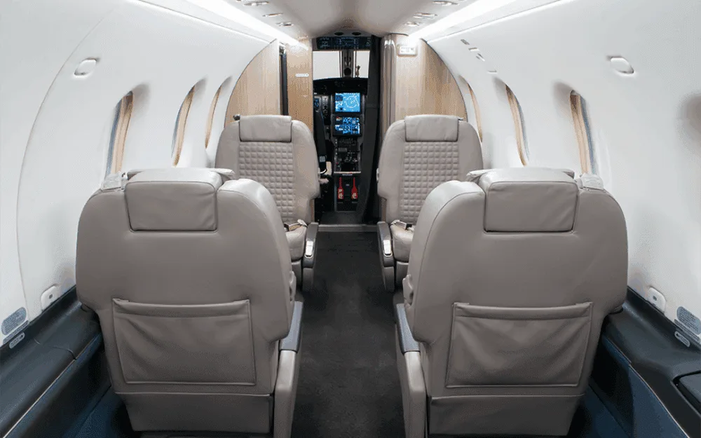 A view down the aircraft cabin aisle shows light grey leather seats and a glimpse of the cockpit beyond.