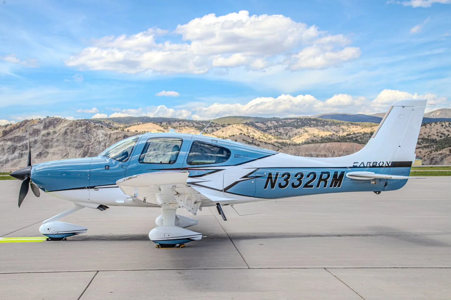 A blue and white Cirrus SR22T GTS aircraft, registration N332RM, parked on an airport tarmac with mountains in the background.