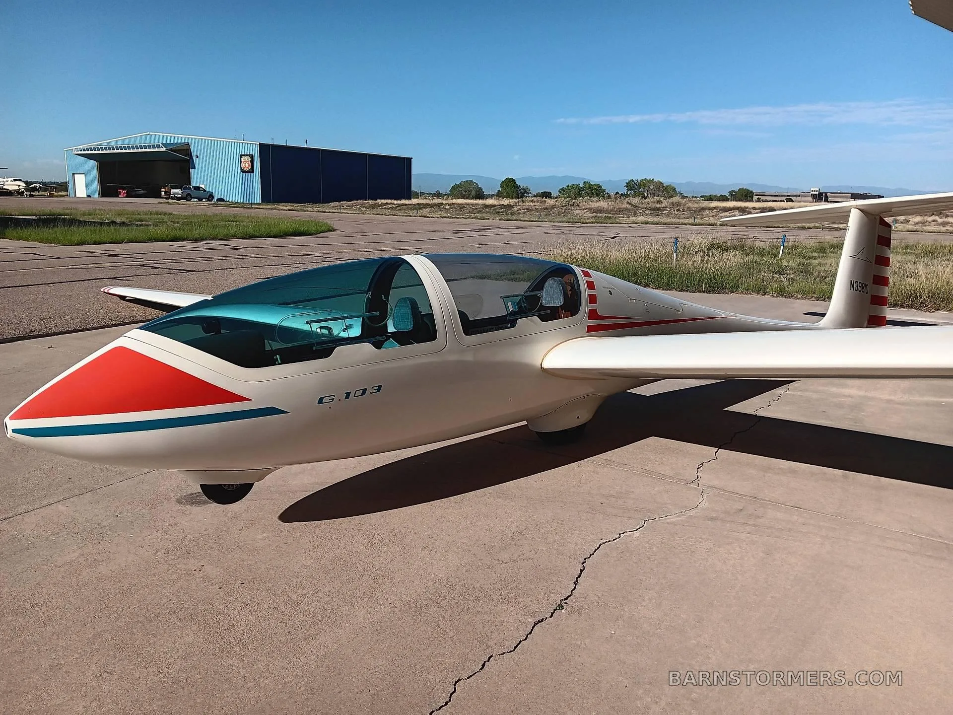 A white Grob G 103 glider with red and blue stripes on the nose, parked on an airfield tarmac with a blue hangar in the background.