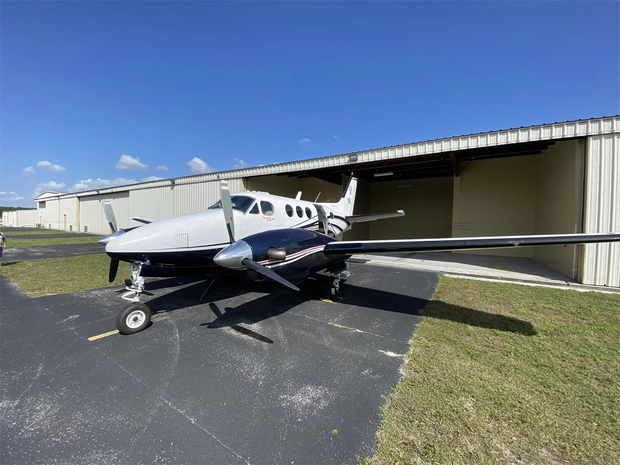 A white and dark blue Beechcraft King Air aircraft parked on an asphalt apron next to a hangar under a clear blue sky.