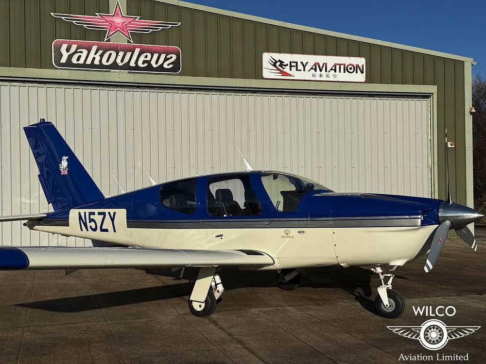 A blue and white single-engine aircraft, N5ZY, parked on an airfield in front of a hangar with 'Yakovlev' and 'Fly Aviation' signs.