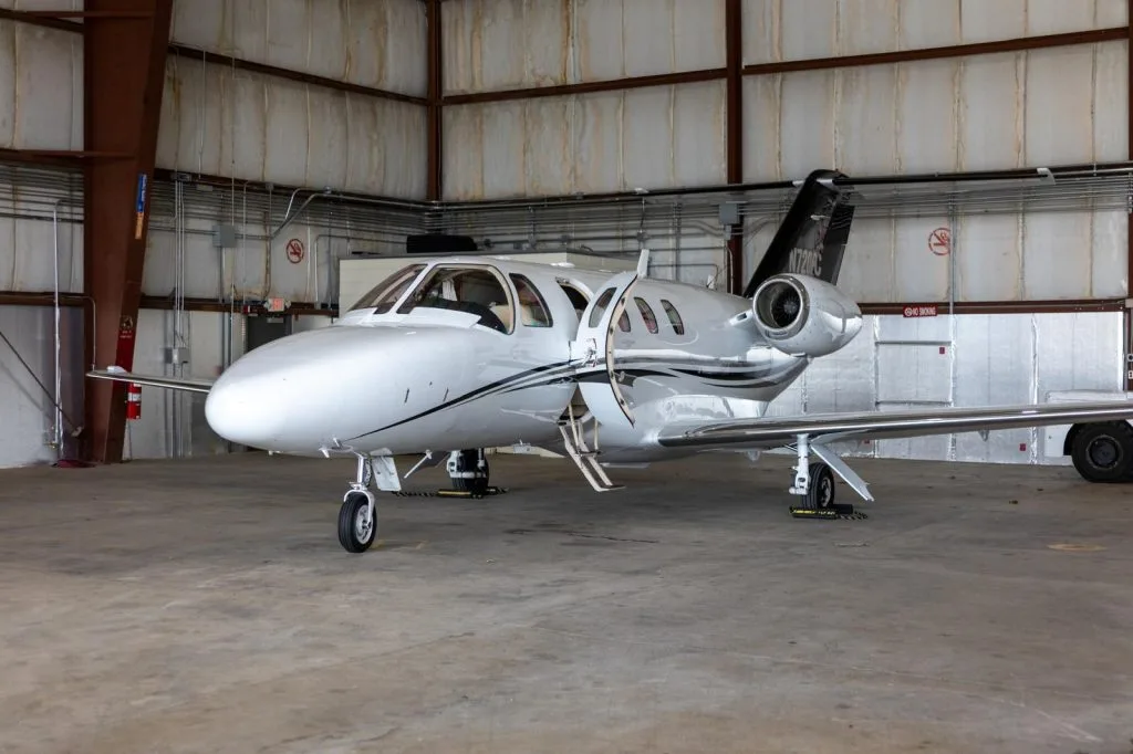 A white Cessna Citation M2 business jet with its main cabin door open is parked inside a hangar.