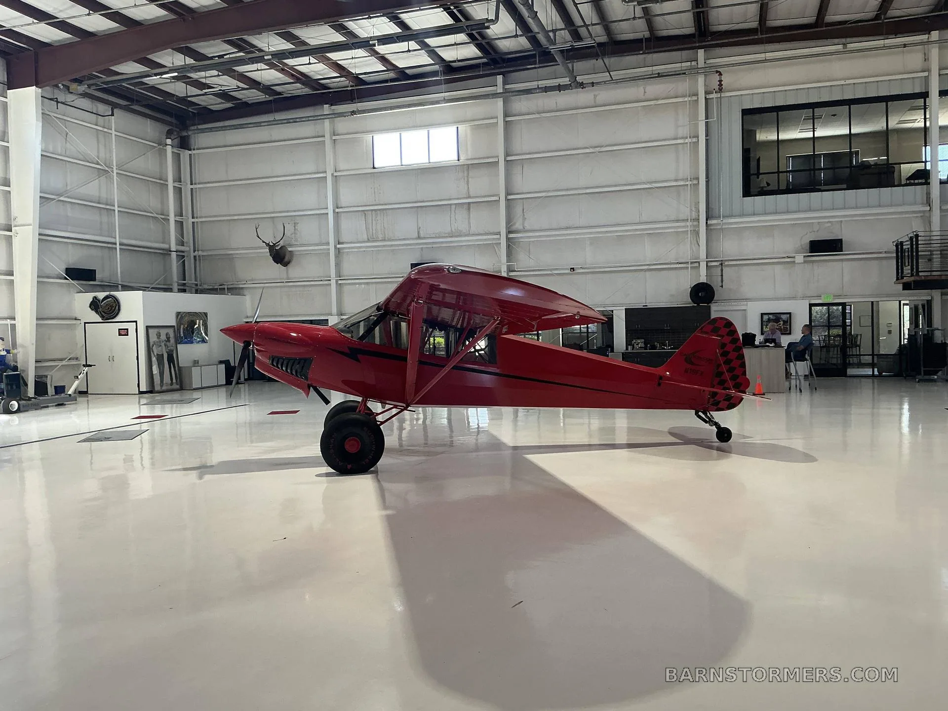 A red Bushmaster aircraft with large tires is parked in a hangar, viewed from the left side.