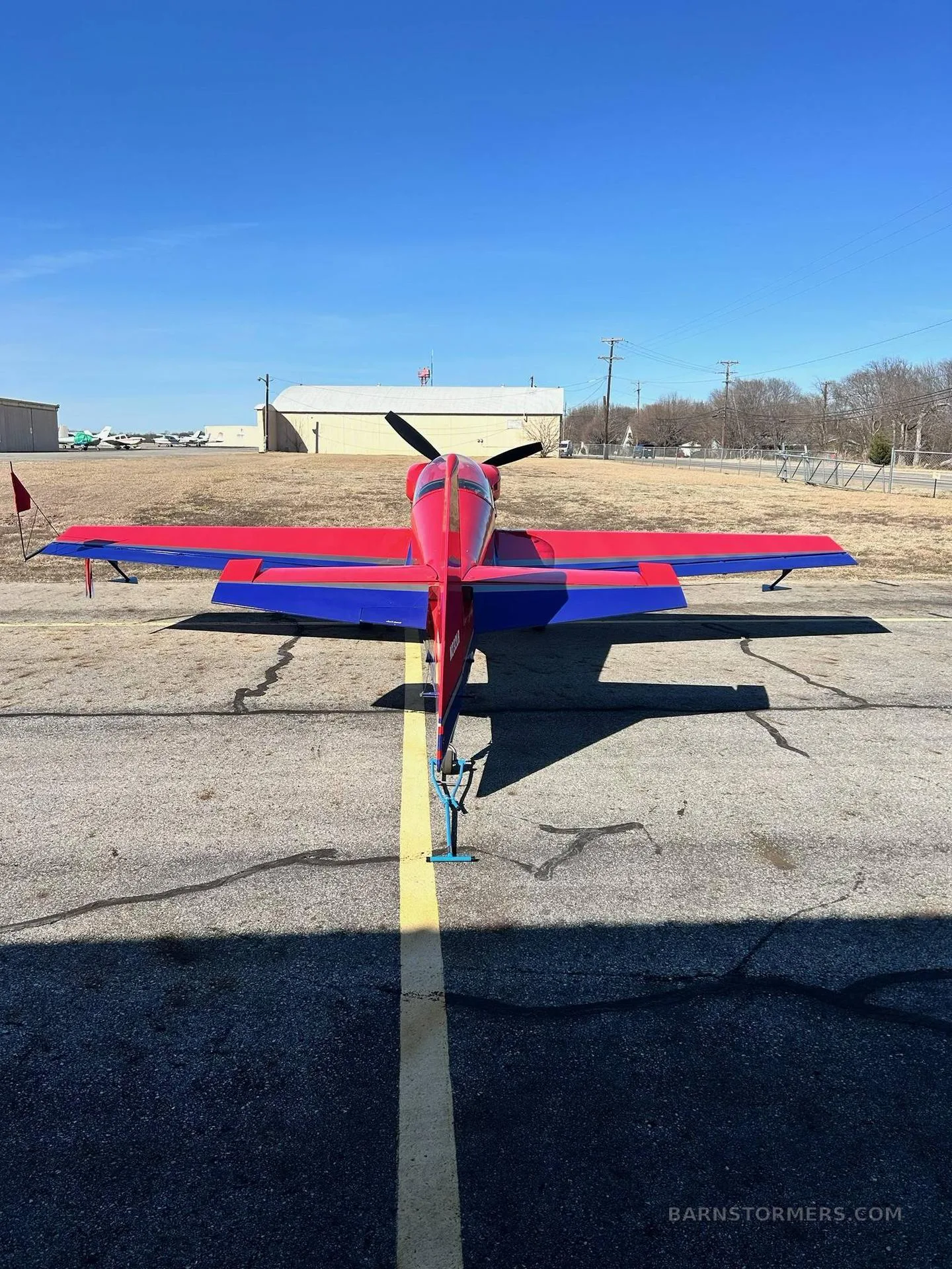 The rear view of a red and blue aerobatic aircraft parked on an asphalt apron with a yellow centerline.