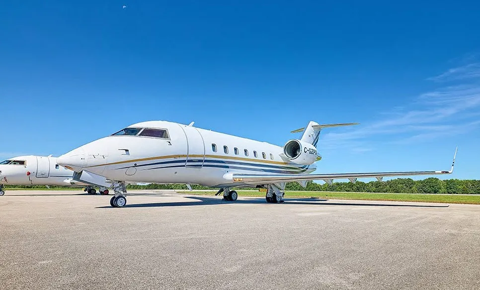 A white Bombardier Challenger business jet with blue and gold stripes is parked on an airport tarmac under a clear blue sky.