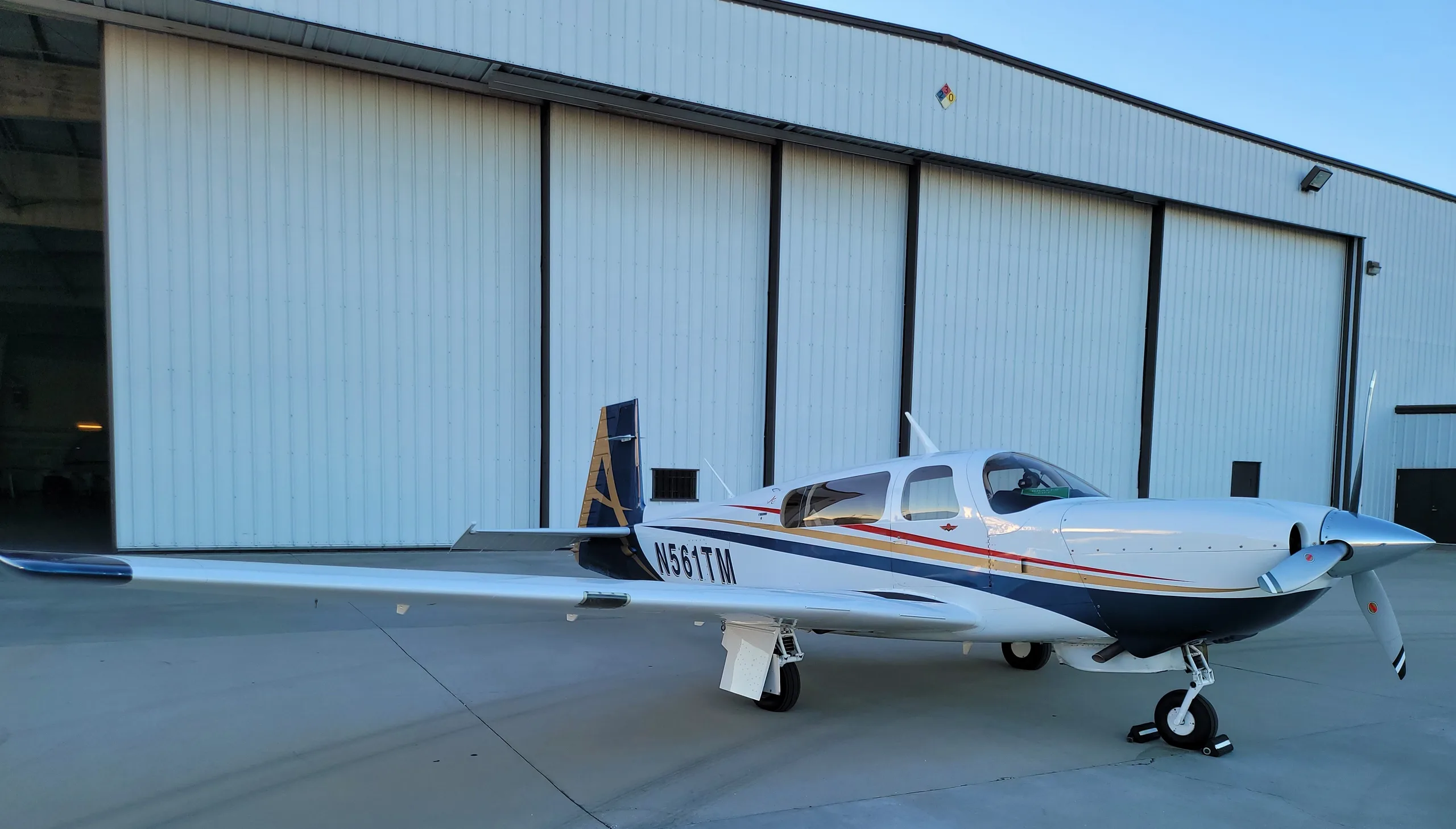 A white Mooney aircraft with blue, gold, and red stripes is parked on a tarmac in front of a white hangar.