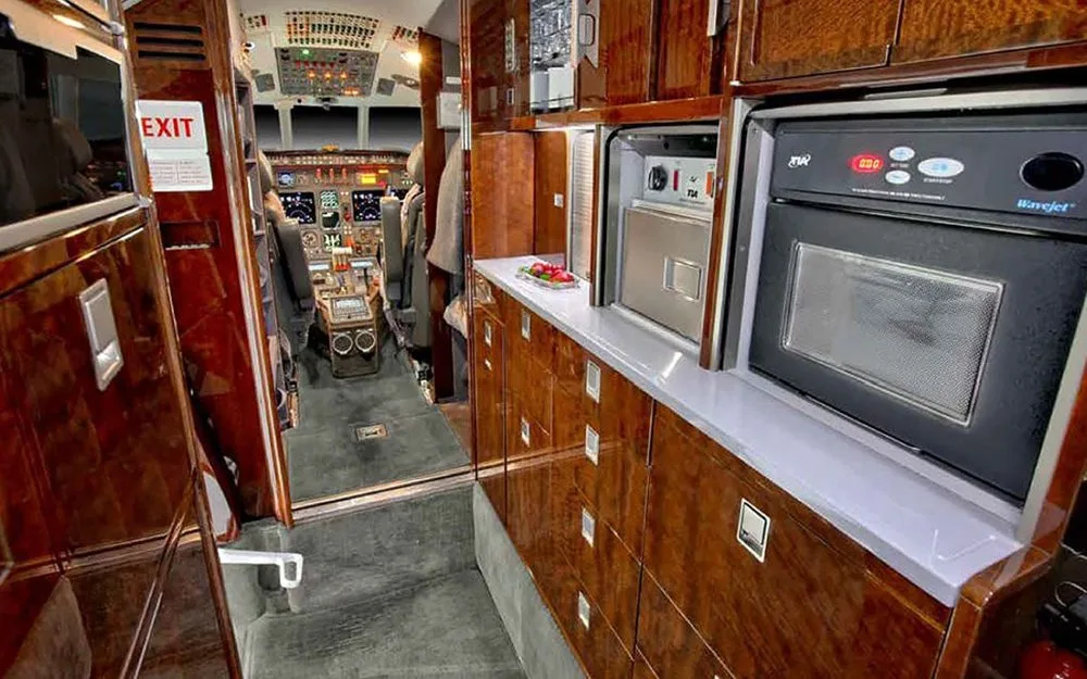The galley area of a business jet with dark wood cabinetry, a microwave, and a view towards the cockpit.