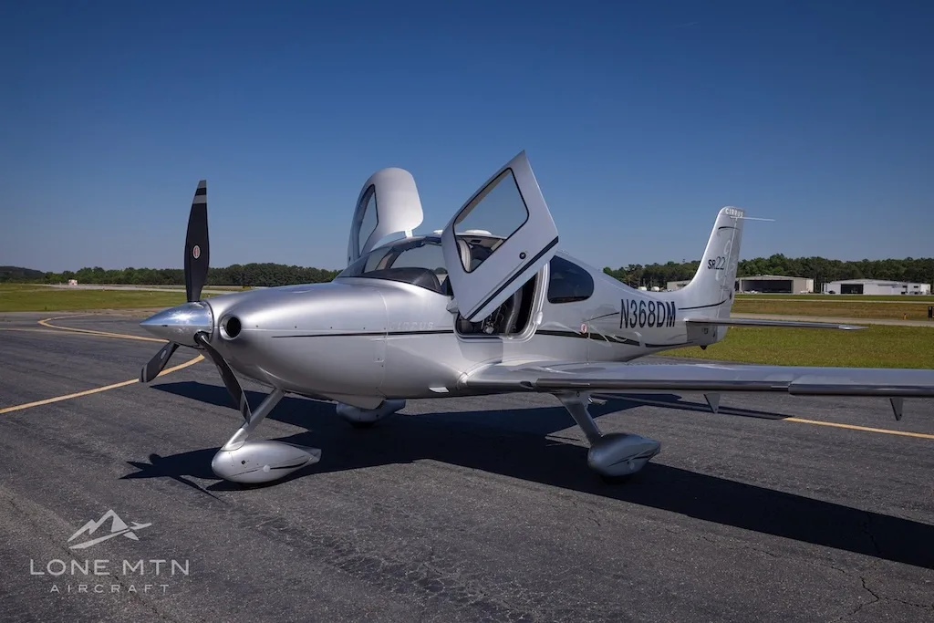 A silver Cirrus SR22 Turbo aircraft (N368DM) with its doors open, parked on an asphalt runway under a clear blue sky.