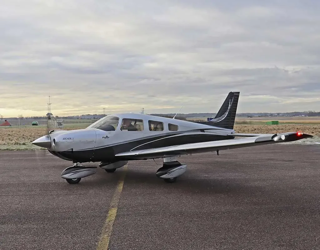 A three-quarter front view of a silver and black Piper Archer LX aircraft on an airfield.