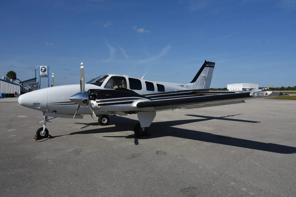 A white and black twin-engine propeller aircraft, N7277G, is parked on an asphalt tarmac with a building in the background.