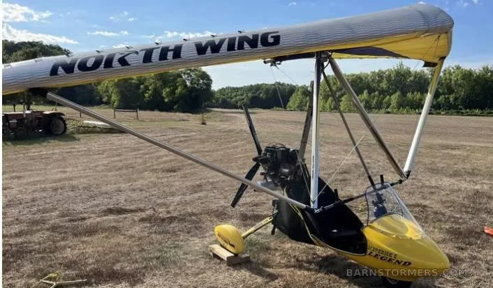 A yellow and silver North Wing Maverick 2 Legend ultralight aircraft sits on a grassy field under a blue sky.
