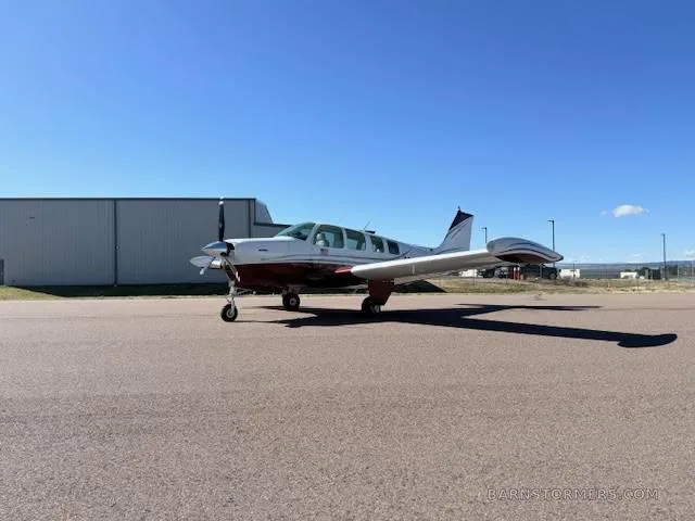 A white and red Beechcraft Bonanza aircraft is parked on an asphalt runway in front of a hangar.