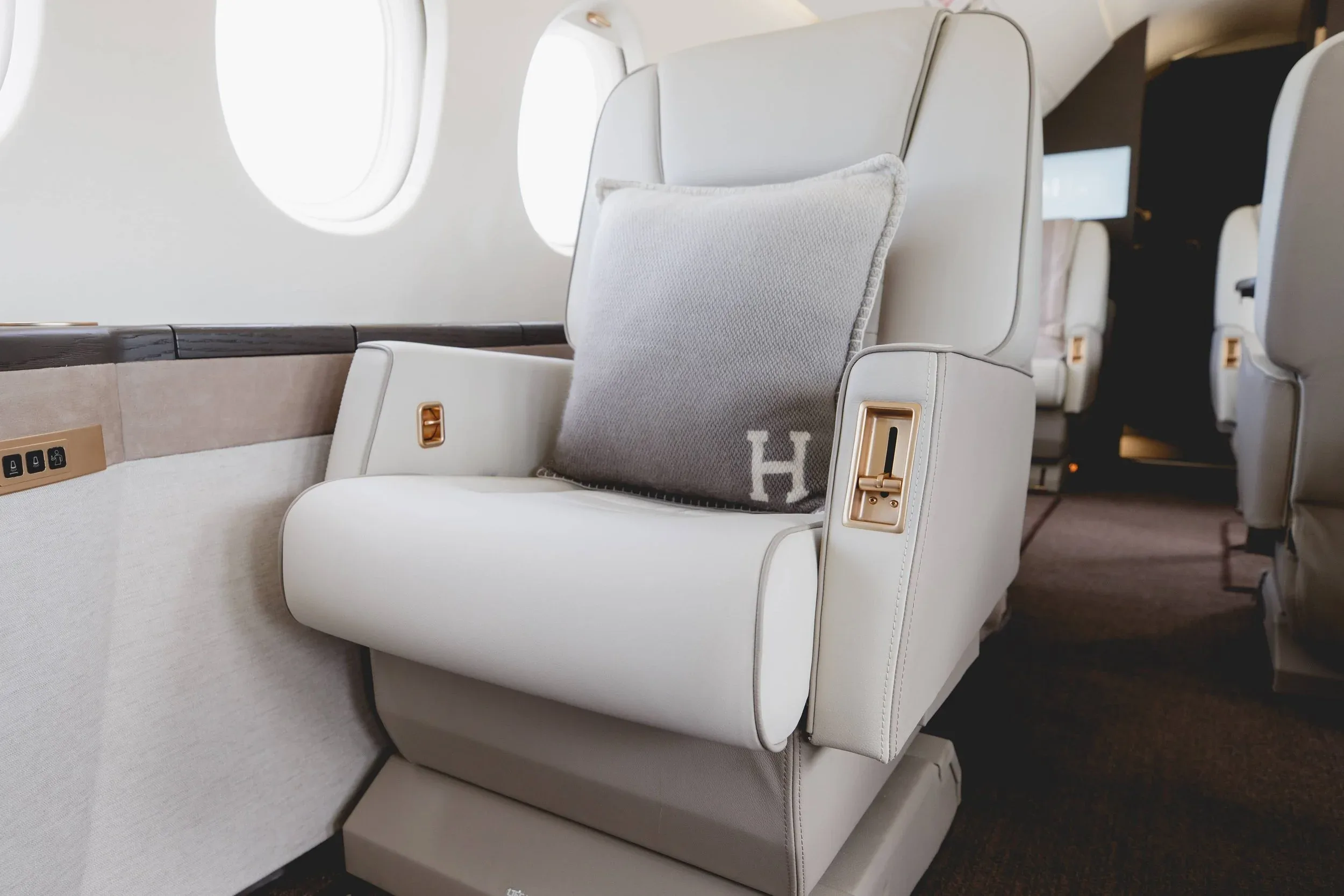 A detailed view of a light-colored leather private jet seat with a grey "H" monogrammed pillow and gold accents.