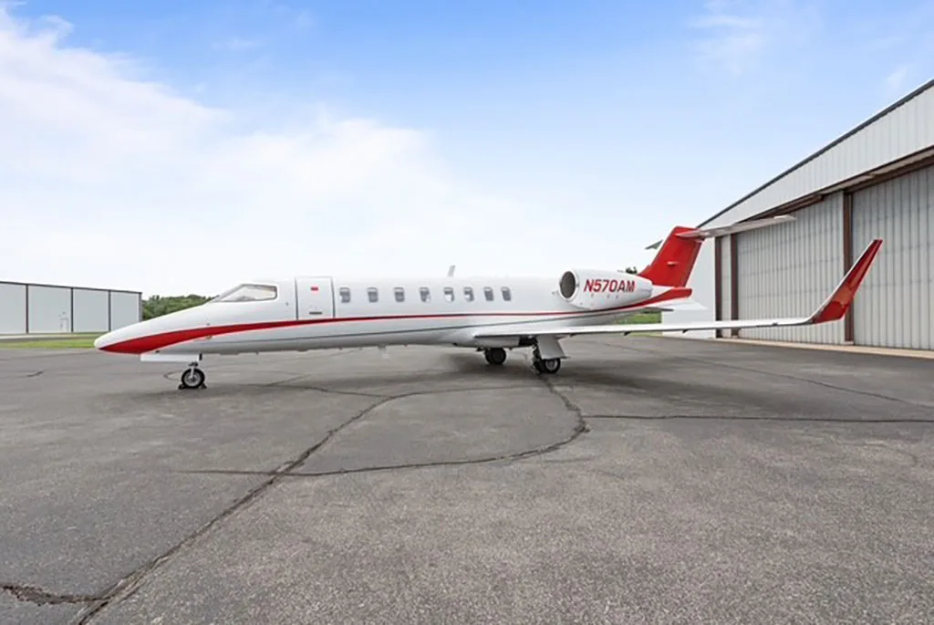 A white and red business jet (N570AM) parked on an asphalt tarmac with a hangar in the background.
