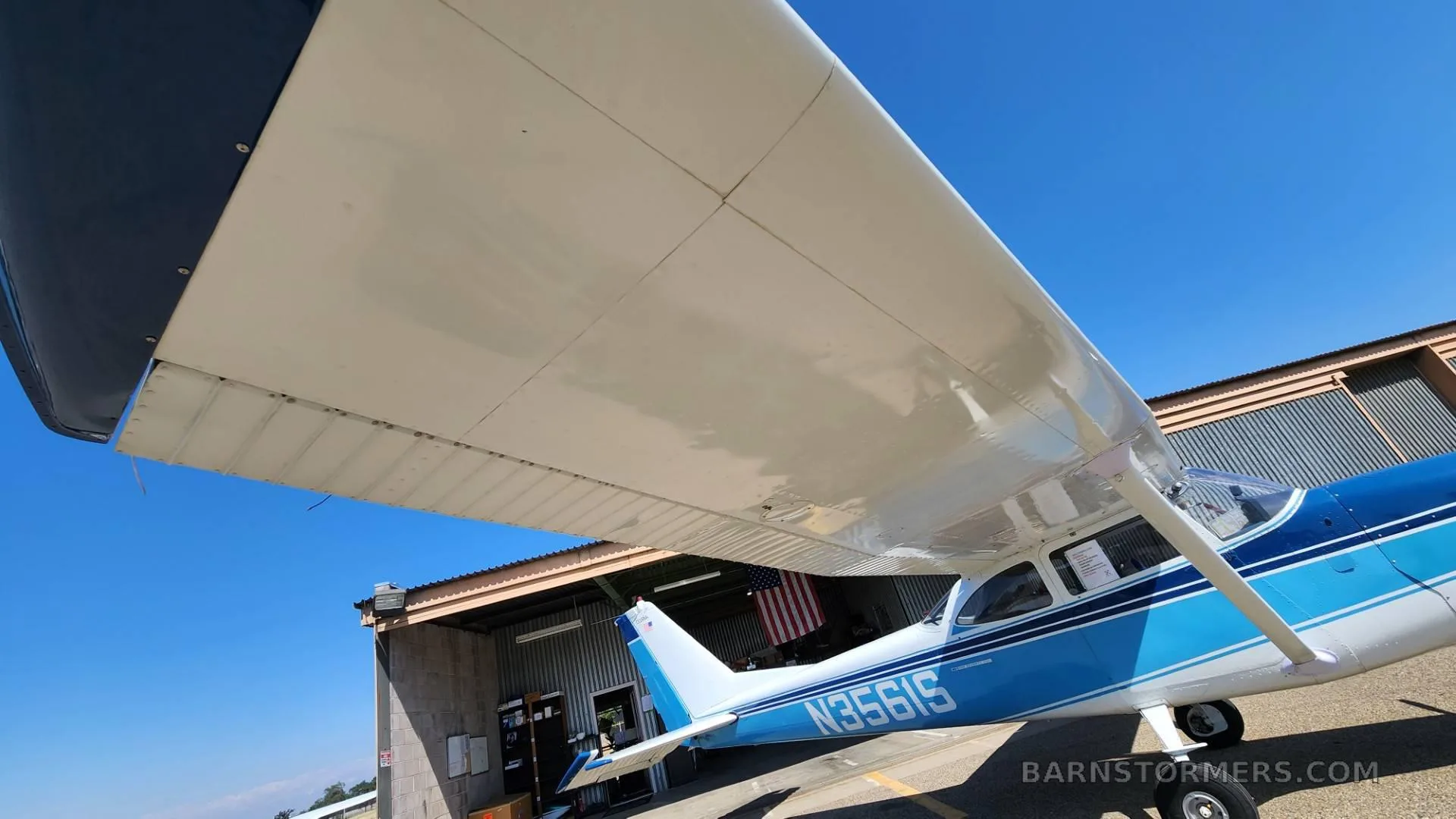 A close-up view of the white wing and blue fuselage of a Cessna aircraft with registration N356IS, parked on an airfield.