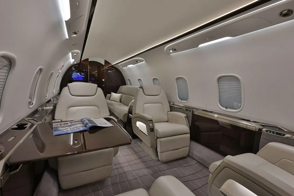 A wide-angle view of the private jet's cabin, showing beige leather seats, a dark wood table with a magazine, and ambient lighting.