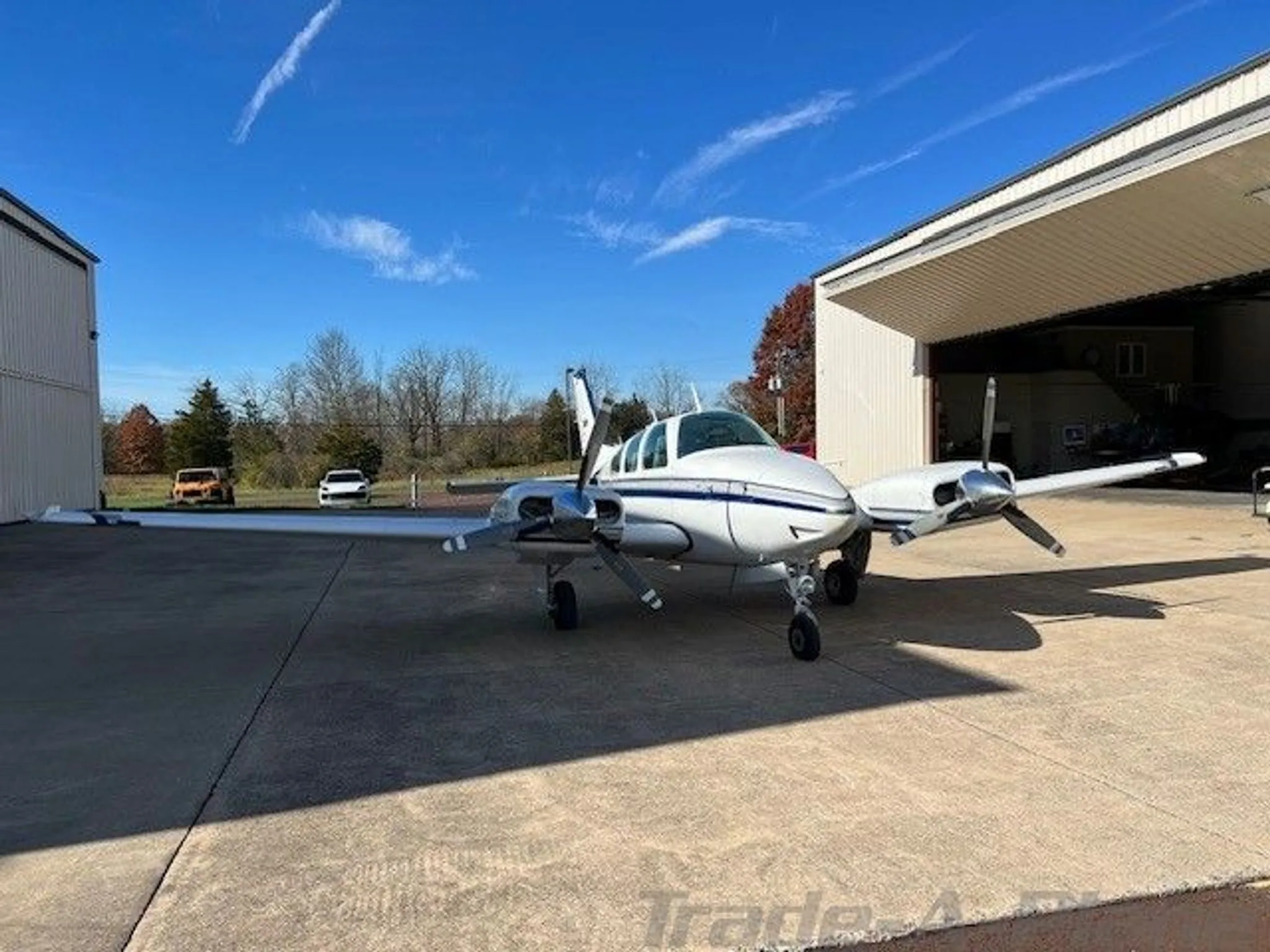 Front-left view of a white twin-engine aircraft with blue and grey stripes parked outside an open hangar.
