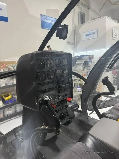 The cockpit of a helicopter shows the instrument panel, controls, and a grey seat.