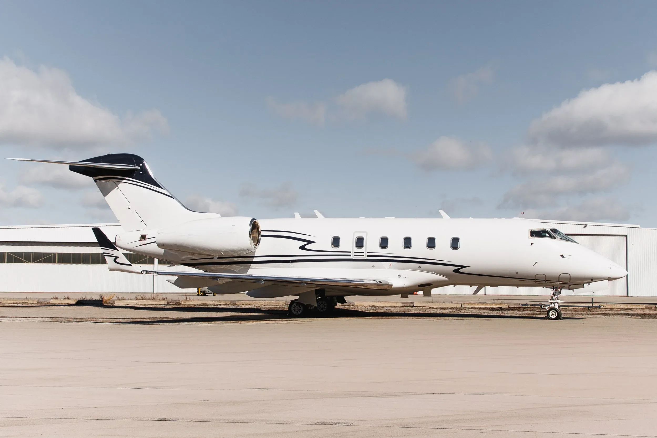 A white business jet with black and blue stripes on the tail and fuselage parked on an tarmac in front of a hangar under a partly cloudy sky.