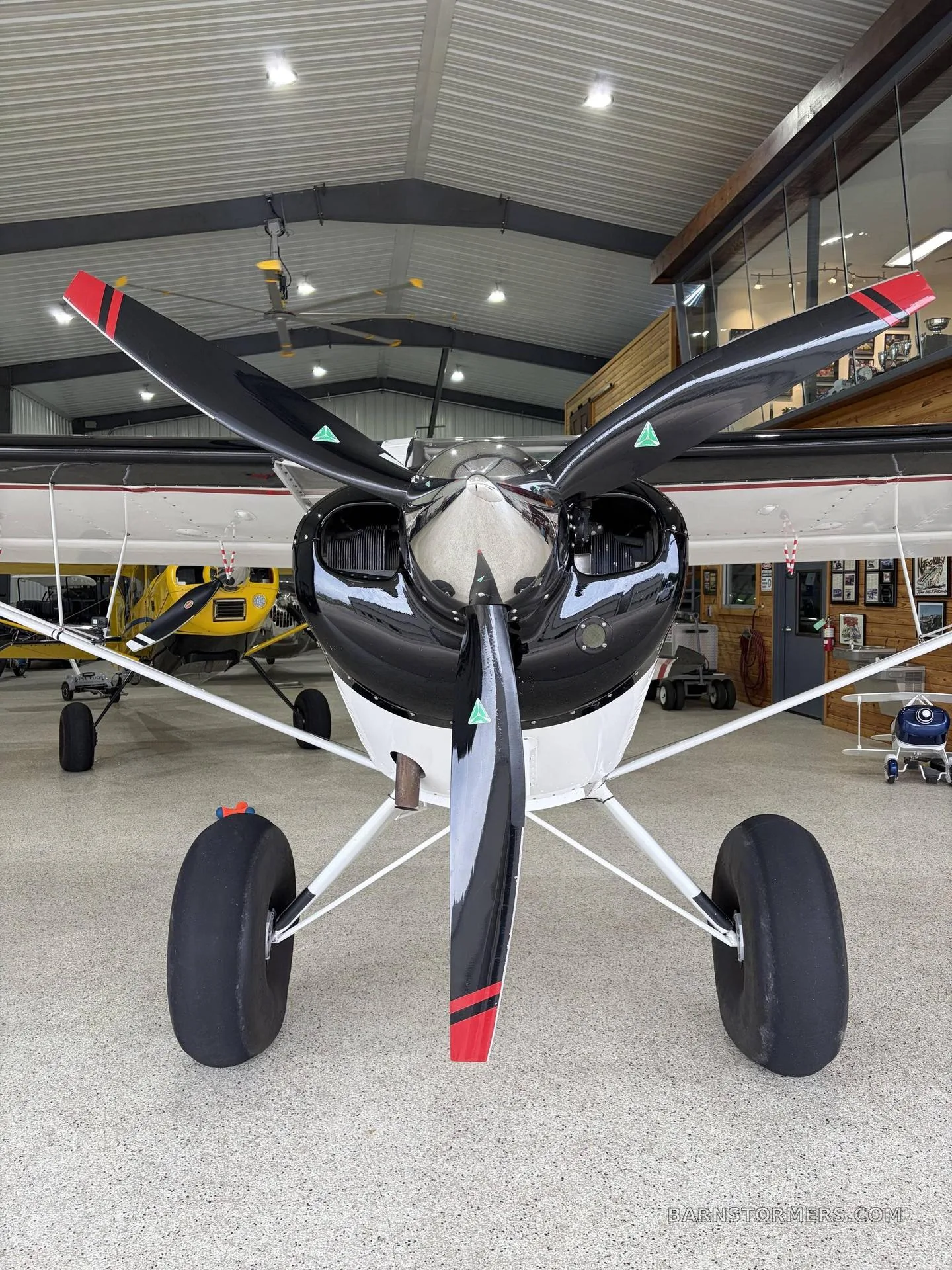 A head-on view of a black and white Aviat Aircraft Husky A-1B with a three-bladed propeller and large tundra tires, parked in a hangar.