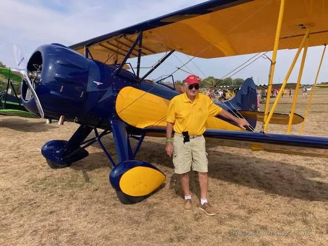 A man in a yellow shirt and red hat stands next to a blue and yellow biplane on a grassy field.