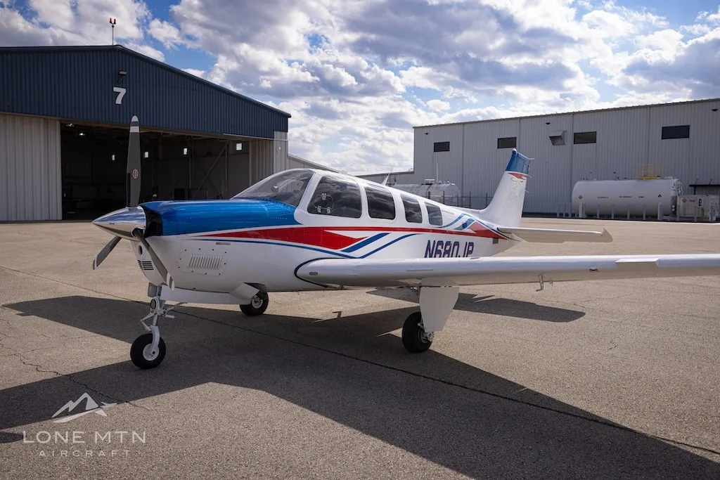 A front-side view of a white, blue, and red Beechcraft Bonanza N680JP parked on an airfield in front of a hangar.