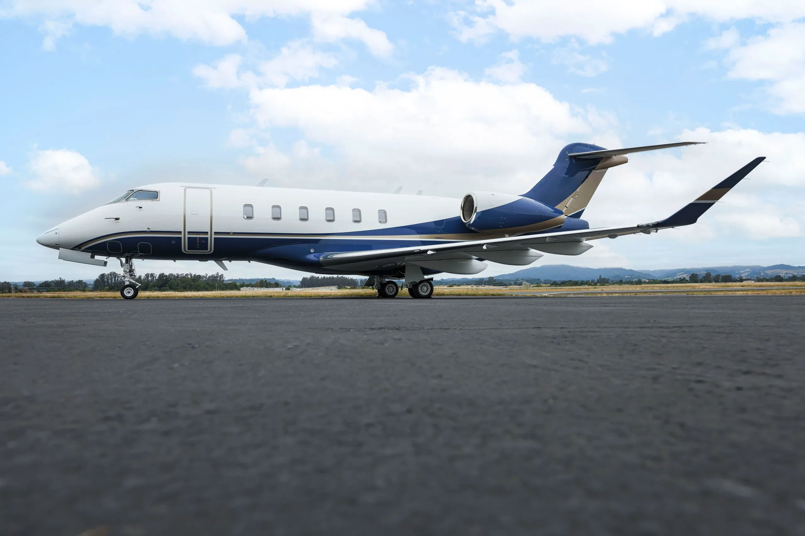 A Bombardier Challenger 350 business jet with a white and blue paint scheme parked on an airport tarmac under a partly cloudy sky.