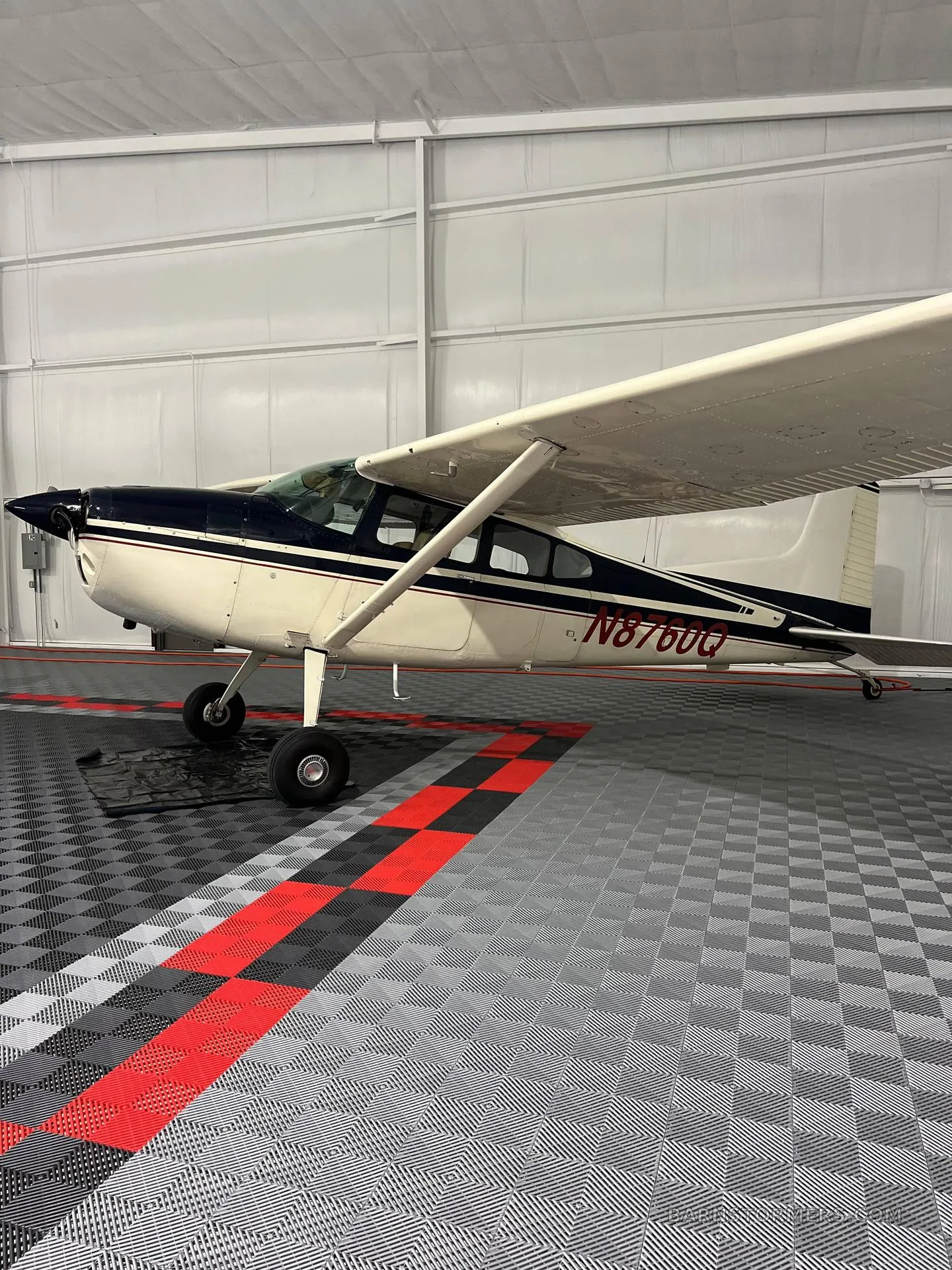 A white and dark blue single-engine high-wing aircraft with registration N876QQ is parked inside a hangar with a gray and red checkered floor.