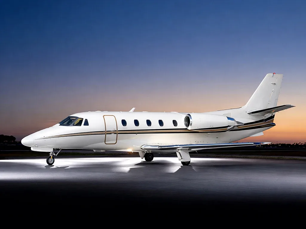 A white business jet with gold and black stripes is parked on a tarmac at dusk, illuminated by ground lights.