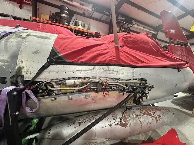 The fuselage of a silver jet aircraft with open access panels revealing internal components, covered by red tarps in a hangar.