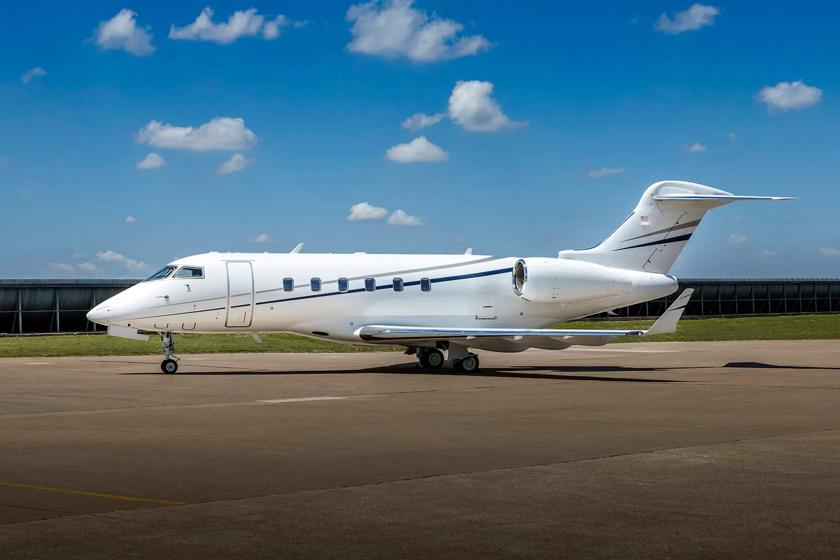 A white business jet with blue and grey stripes is parked on an airfield under a blue sky with scattered clouds.