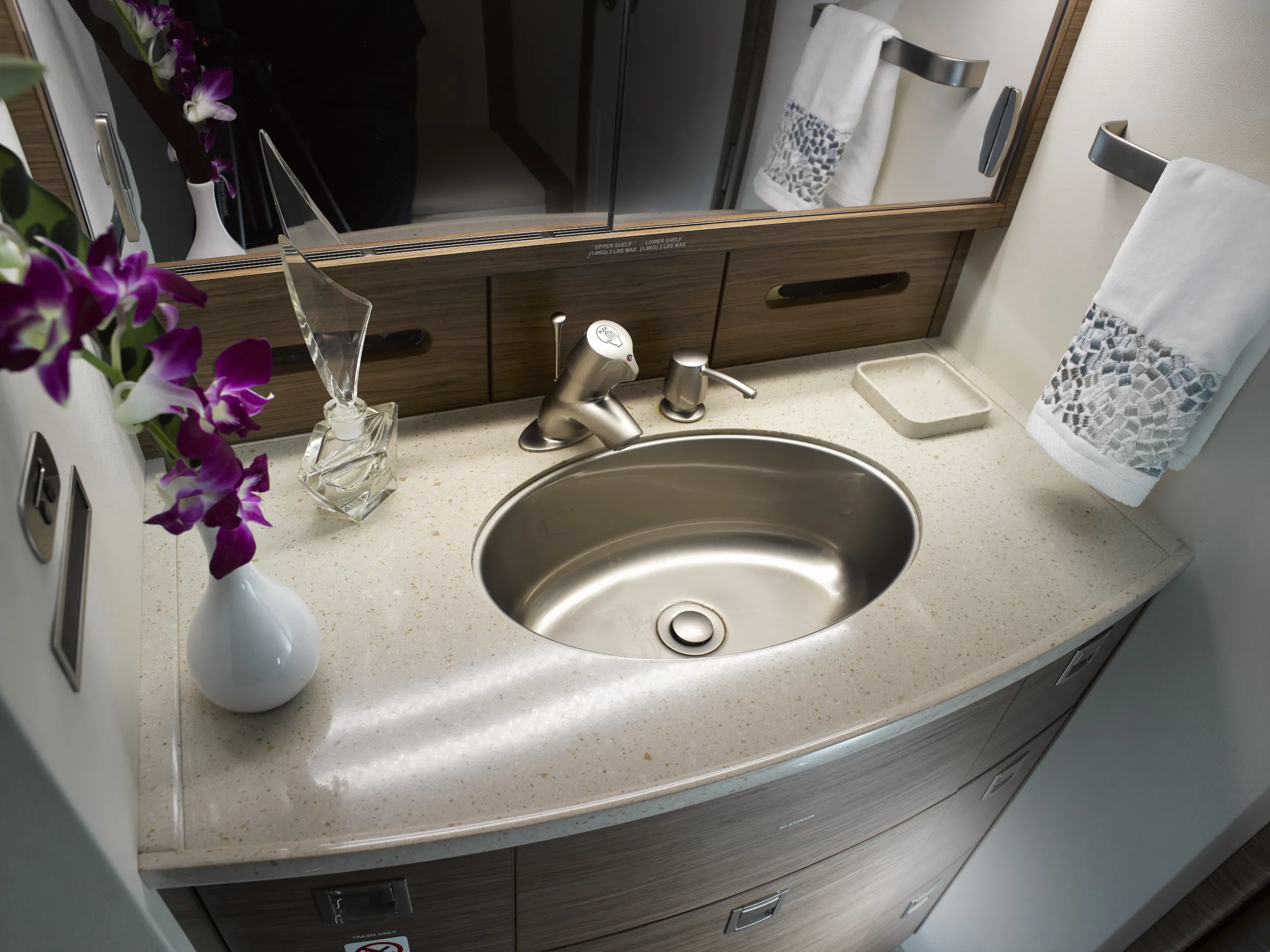 A modern aircraft lavatory sink with a polished faucet, a light-colored countertop, and purple orchids in a vase.