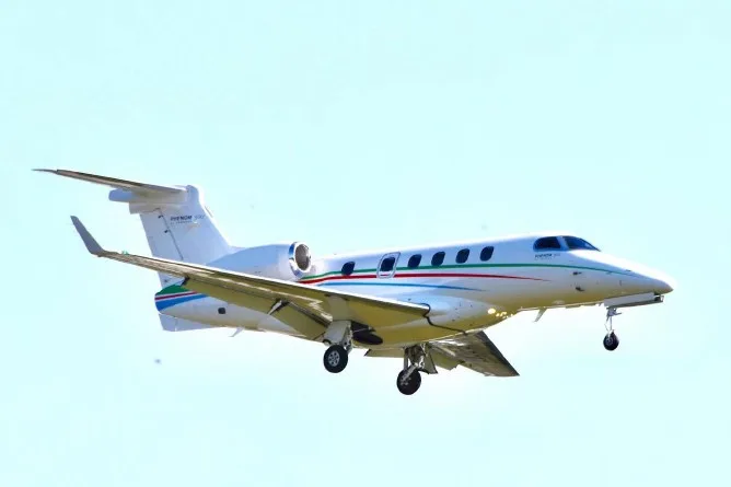 A white Embraer Phenom 300 business jet with green and red stripes flies against a clear sky with its landing gear extended.