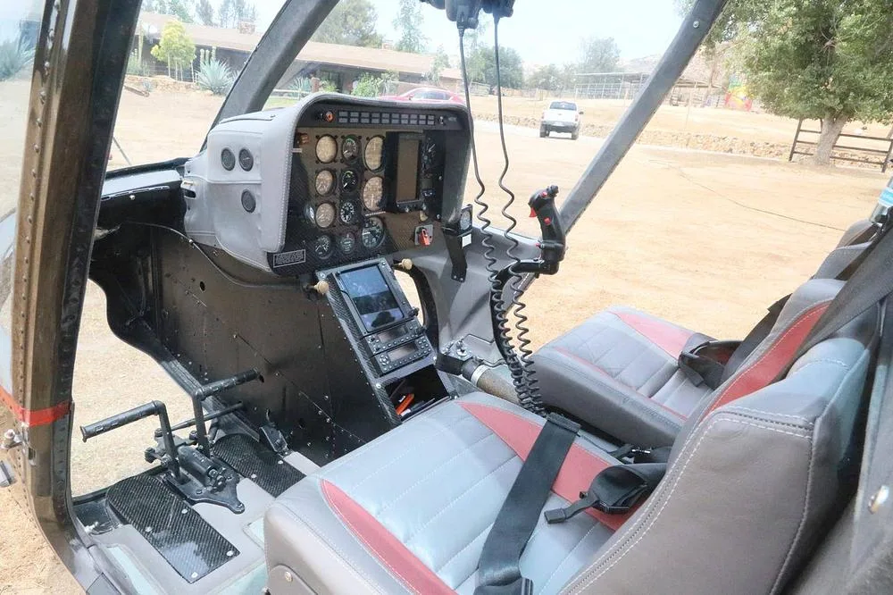 The cockpit of a helicopter shows the instrument panel, controls, and two grey and red seats.