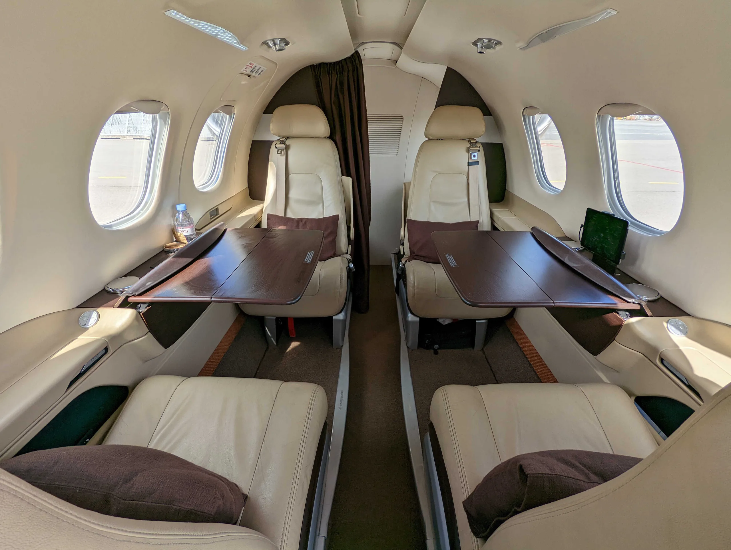 The interior cabin of a private jet features four beige leather seats with dark brown pillows and two fold-out tables.