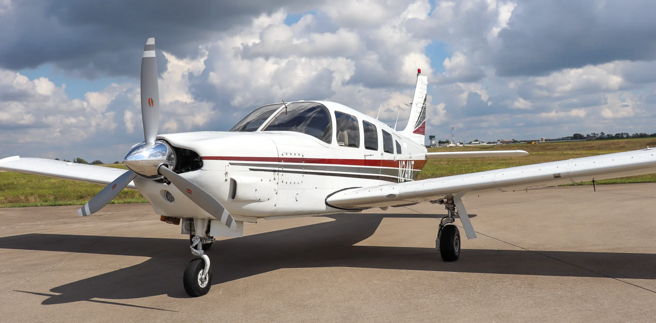 A white Piper Lance (N24MT) with red and grey stripes is parked on an airfield, viewed from the front-left.