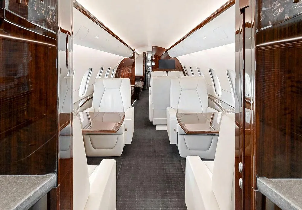 View down the aisle of a private jet cabin featuring cream seats, dark wood paneling, and a dark carpet.