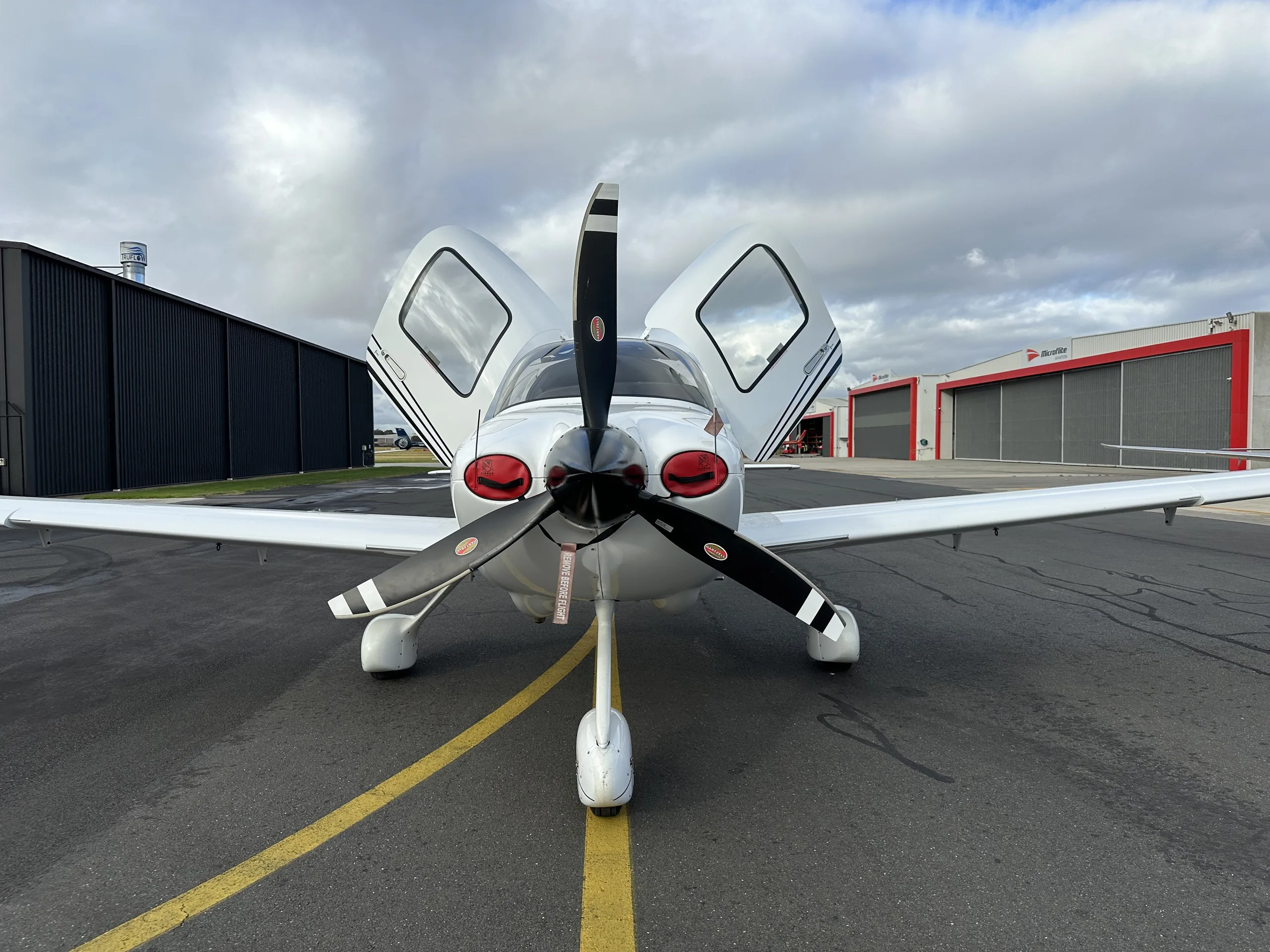 A front view of a white Cirrus SR22 aircraft with its gull-wing doors open, parked on an airfield.