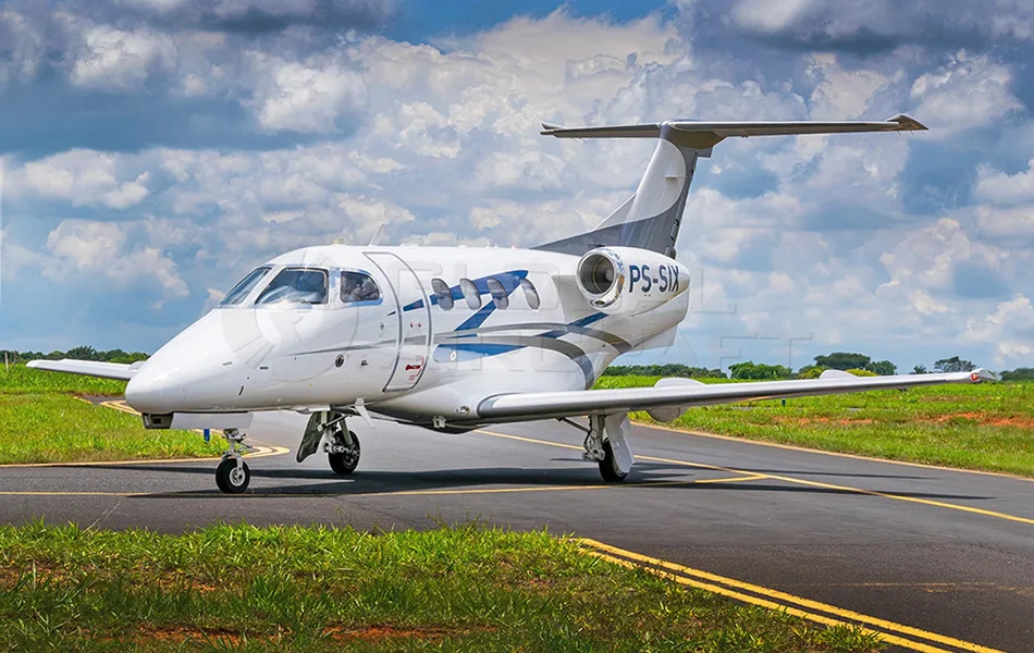 A white Embraer Phenom 300 private jet with blue and grey stripes is parked on a tarmac with green grass under a cloudy sky.