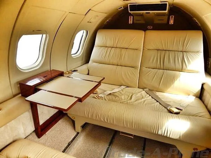The aircraft cabin interior features light beige leather seating, a fold-out table, and large windows.